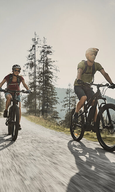 zwei Personen fahren auf E-Bikes auf einem Schotterweg, umgeben von einer Berglandschaft 
