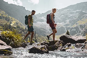 Zwei Wanderer überqueren einen Bach in einer alpinen Landschaft mit Bergpanorama in Schladming