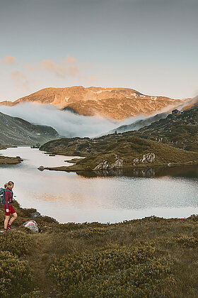 Zwei Wanderer laufen am Giglachsee entlang, umgeben von majestätischen Bergen bei Sonnenaufgang