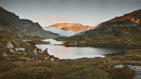 Zwei Wanderer laufen am Giglachsee entlang, umgeben von majestätischen Bergen bei Sonnenaufgang