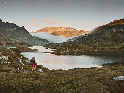 Zwei Wanderer laufen am Giglachsee entlang, umgeben von majestätischen Bergen bei Sonnenaufgang