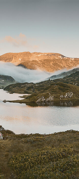 Zwei Wanderer laufen am Giglachsee entlang, umgeben von majestätischen Bergen bei Sonnenaufgang